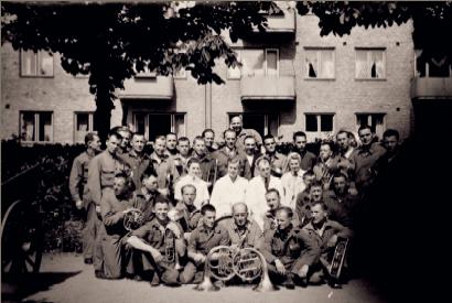 A black and white picture of a group of former Polish prisoners standing in front of a house in Malmö/Sweden, some of them holding musical instruments in their hands.