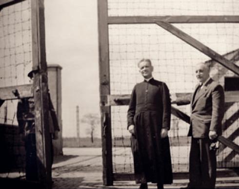 Abbot Noël Carlotti and another man stand in front of an open barb wire fence gate of Neuengamme concentration camp and look into the camera.
